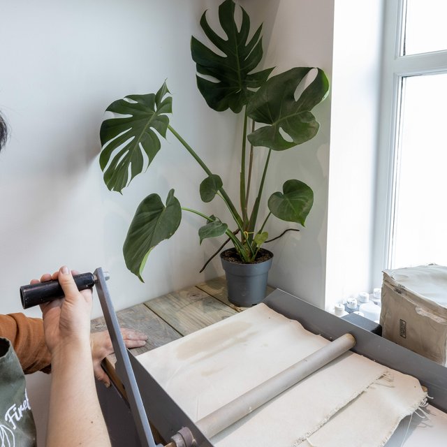 A person operates a printing press near a potted monstera plant. A cardboard box sits nearby. The scene is brightly lit by a window.
