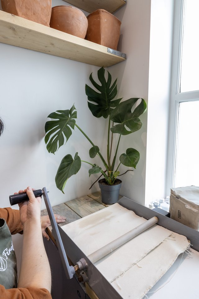 A Monstera plant sits in a black pot on a wooden shelf near a window. A person's hands and a roll of fabric are visible in the foreground.