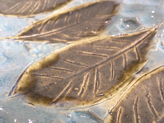 A textured, bronze-colored leaf sits atop a textured, pale blue surface. Details of the leaf's veins are visible.