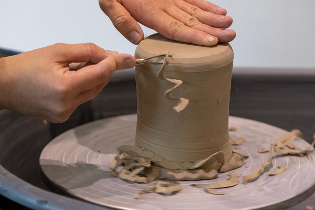 A potter's hands shape a cylindrical clay form on a pottery wheel. Clay scraps are scattered around the wheel.