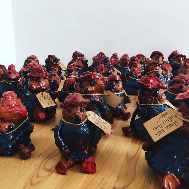 A large group of small, reddish-brown clay figures wearing blue hats and tagged with handwritten labels are arranged on a light-colored wood floor.