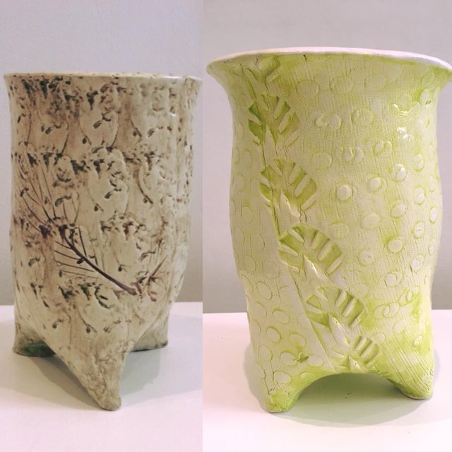 Two ceramic planters sit on a white surface. One is beige with brown floral designs, the other is light green with leaf patterns. Both have a textured surface and three legs.