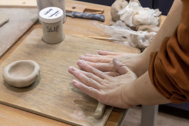 A person's hands are shaping a ball of clay on a wooden table. A jar labeled 'SLIP' and a partially formed clay bowl are nearby.
