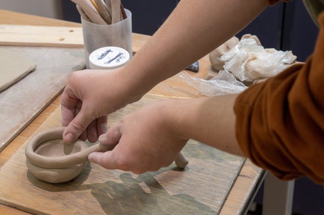 A person's hands mold a coil of gray clay on a wooden table. A jar of slip and a ball of twine are nearby.