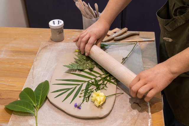A person presses leaves and flowers into a sheet of gray clay on a wooden table. Tools and a jar of slip are nearby.