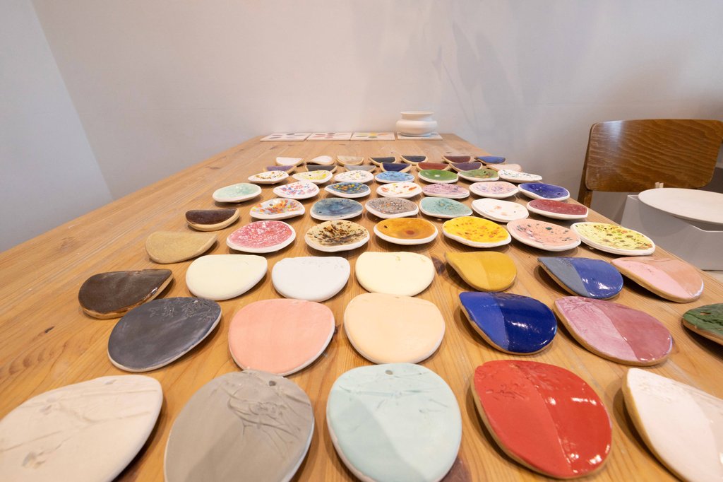 Numerous small, colorful ceramic dishes are arranged on a wooden table. A wooden chair sits in the background.
