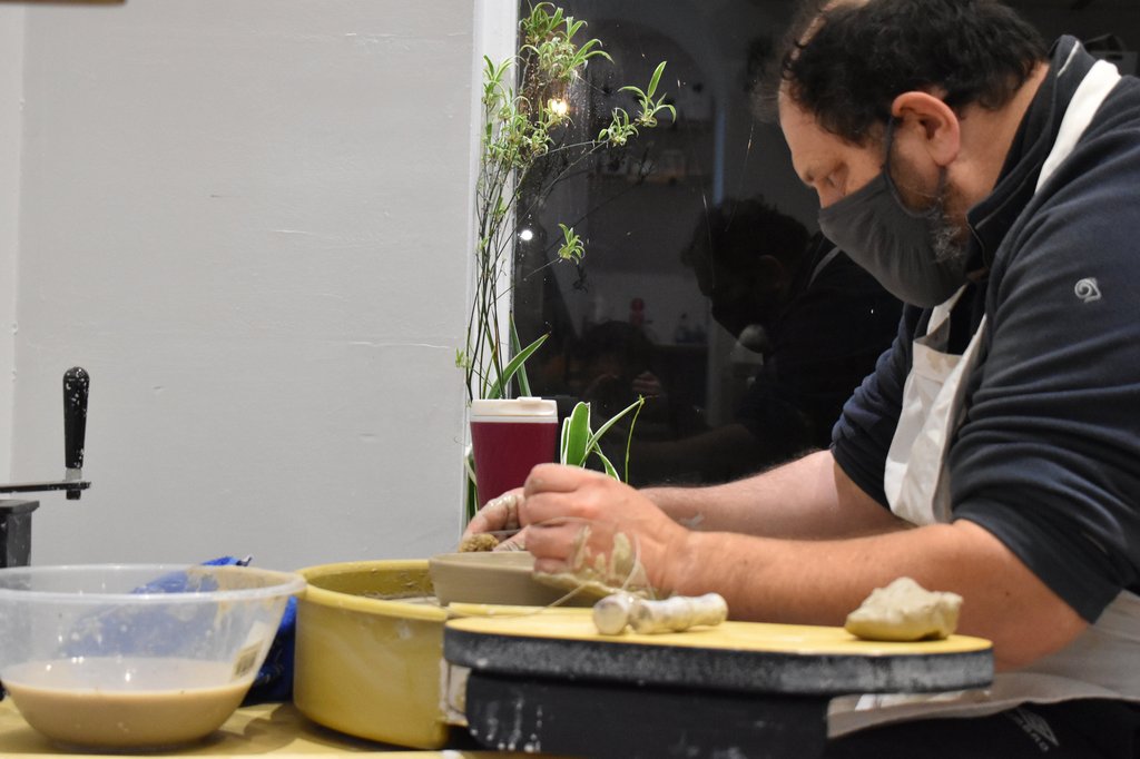 A man in a mask prepares food, possibly dumplings, on a wooden surface. A bowl of dough and a small plant are visible in the background.