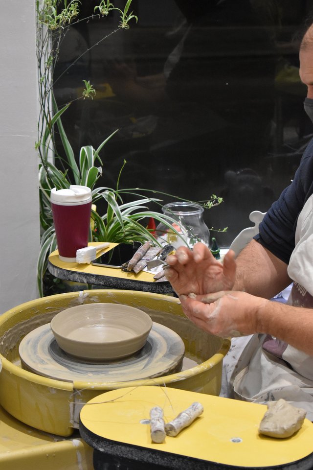 A person's hands shape clay on a pottery wheel. A cup of coffee and plants sit on a nearby table. The background is dark and out of focus.