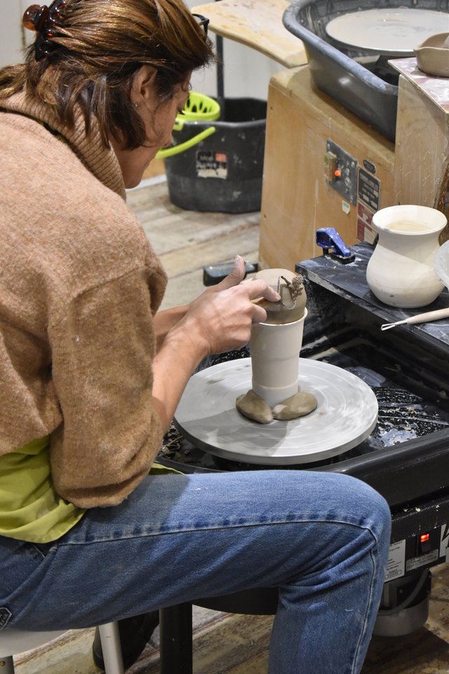 A person sits at a pottery wheel shaping clay. A finished vase sits nearby. The background includes a dark bucket and some tools.