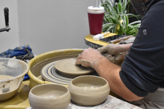 Close-up of hands shaping clay on a pottery wheel. Two unfinished bowls sit on the wheel alongside a partially formed piece. A red cup and plants are visible in the background.