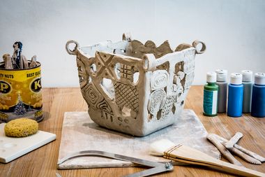A textured, gray clay sculpture sits on a wooden surface. Tools and paints are nearby. A bag of nuts and a sponge are also visible.