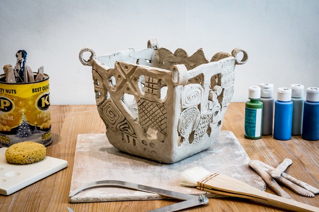 A textured, gray clay sculpture sits on a wooden surface. Tools and paints are nearby. A bag of nuts and a sponge are also visible.