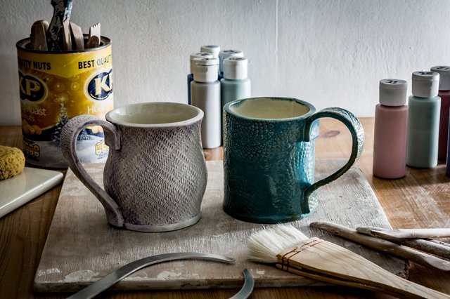 Two textured ceramic mugs sit on a wooden workbench covered with a linen cloth. Brushes, paints, and food containers are nearby.