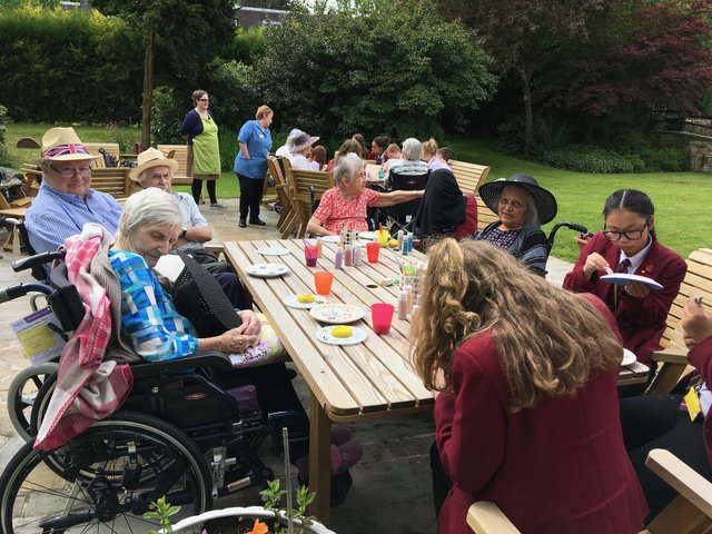 People of various ages sit at picnic tables outdoors. Some are in wheelchairs, others are standing or seated. A woman in a red uniform is eating.