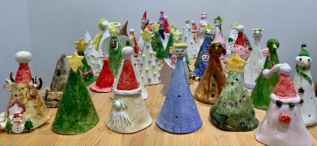 A collection of ceramic figures arranged on a wooden table. The figures are predominantly cone-shaped, decorated with holiday themes like snowmen, Christmas trees, and Santa hats.