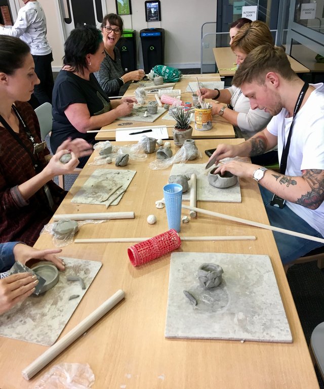 Several people are seated at a long table, sculpting clay on tiles. Tools and supplies are scattered across the workspace.