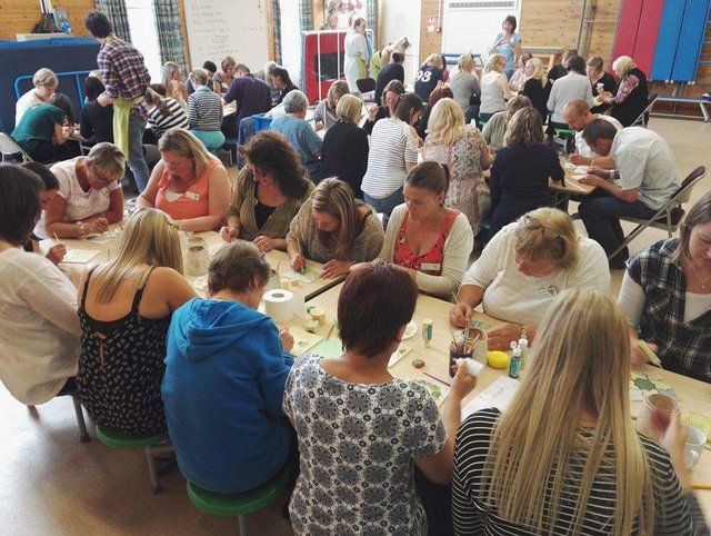 A large group of people are gathered around tables with craft supplies. They appear to be engaged in an activity or workshop.