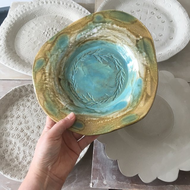 Ceramic bowl with turquoise and brown glaze, held by a hand. White plates in the background.
