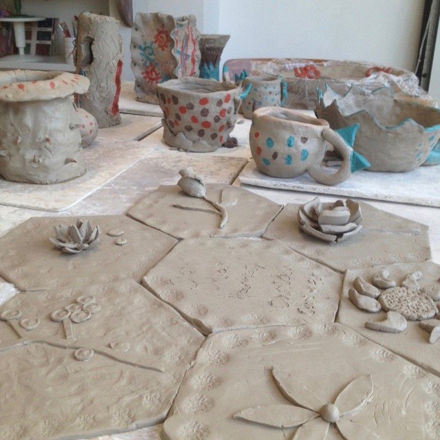 A collection of clay pots and floral sculptures on a textured surface. Some pots have blue and red details, while others are plain. A few clay flowers are scattered around.