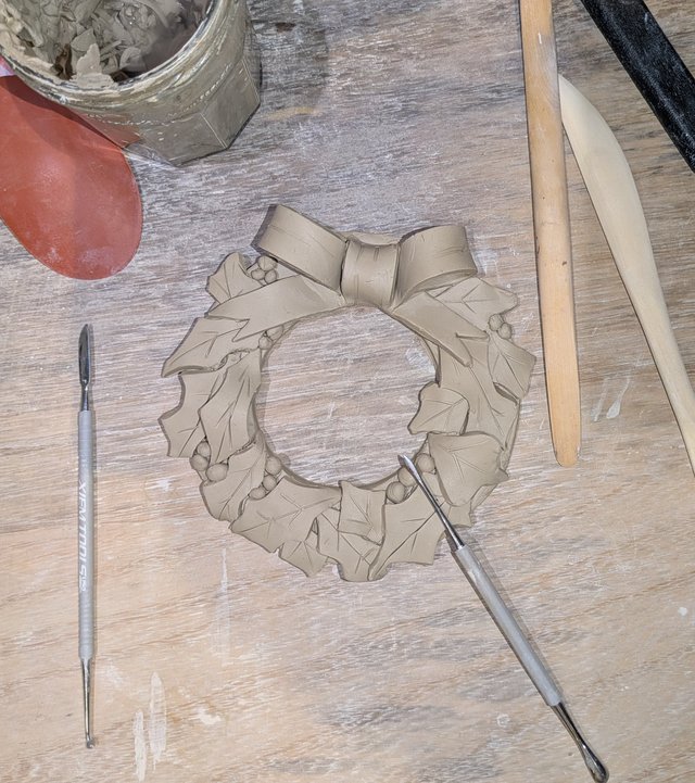 A close-up view of a handcrafted clay wreath in progress, lying on a textured wooden surface.  Tools and clay materials surround the wreath, suggesting an artistic creation process. The wreath has a ruffled, layered design.