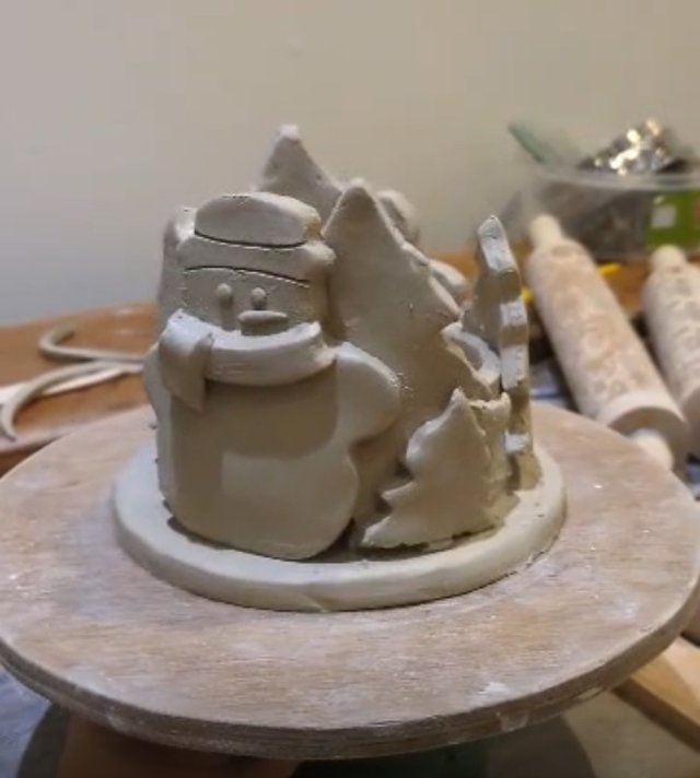 A work-in-progress clay sculpture of Santa Claus emerging from a forest of pine trees, mounted on a pottery wheel base. The sculpture has a rough, unglazed texture, suggesting it's a handmade piece in the early stages of creation.